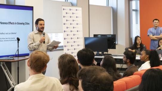 A professor speaking into a microphone in front of a screen facing an audience of students.